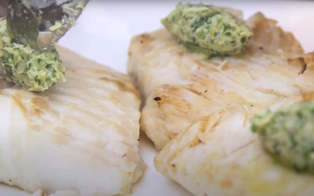 Generate an image of a grilled halibut fillet topped with a compound garlic thyme butter, served with lemon wedges on a warmed platter. baked halibut recipes