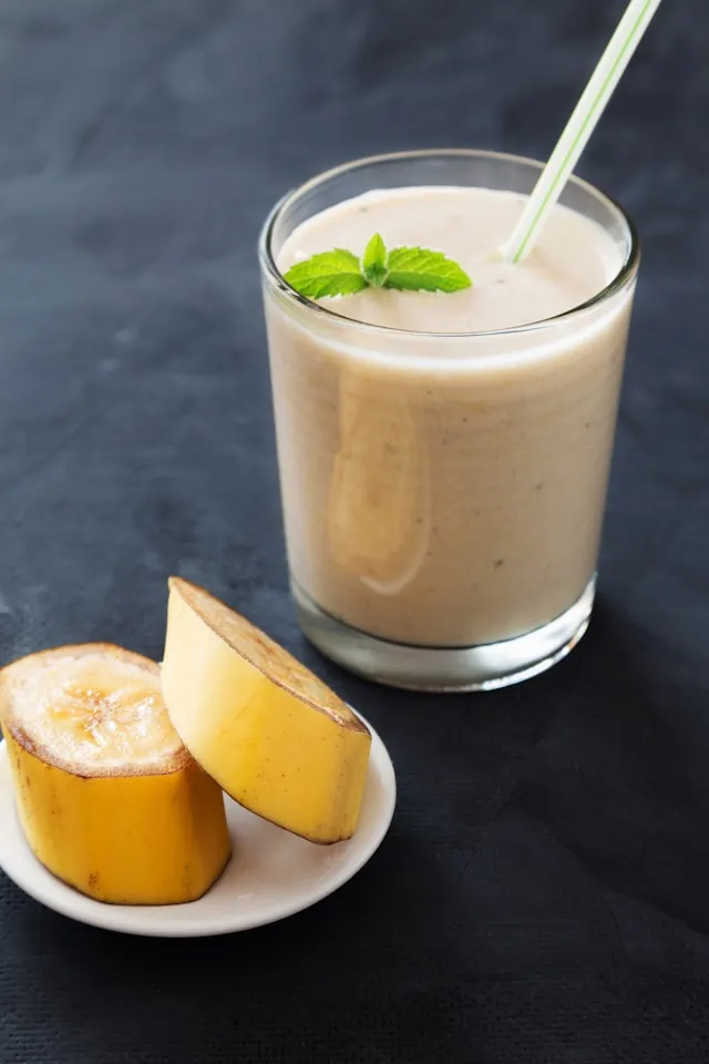 A thick, creamy banana smoothie in a clear glass, with a vibrant yellow-orange hue and a luscious, velvety texture. Sunlight streams through the glass, creating a warm, inviting atmosphere. The smoothie is topped with a dollop of fresh whipped cream, gently swirled and sprinkled with a dusting of ground cinnamon. Ripe banana slices are artfully arranged around the rim of the glass, adding a natural, organic touch. The overall composition is balanced, with a focus on the smoothie as the central subject, showcasing its tempting appearance and inviting the viewer to enjoy a delightful, nourishing breakfast. Image of a classic banana smoothie in a tall glass with a sprinkle