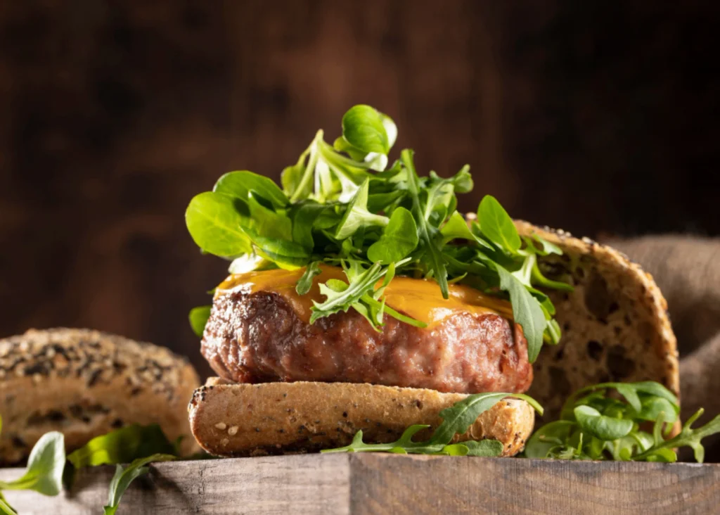A closeup shot of a deer meat burger with melted cheese, tomato, lettuce, and onion, resting on a wooden board. The burger is garnished with fresh sprigs of thyme and rosemary, casting warm, natural shadows. The lighting is soft and diffused, highlighting the juicy texture of the meat and the vibrant colors of the toppings. The background is blurred, creating a focus on the delectable burger in the foreground. The overall mood is appetizing and inviting, showcasing the hearty, comforting qualities of this homemade deer meat recipe. Deer Meat Burgers