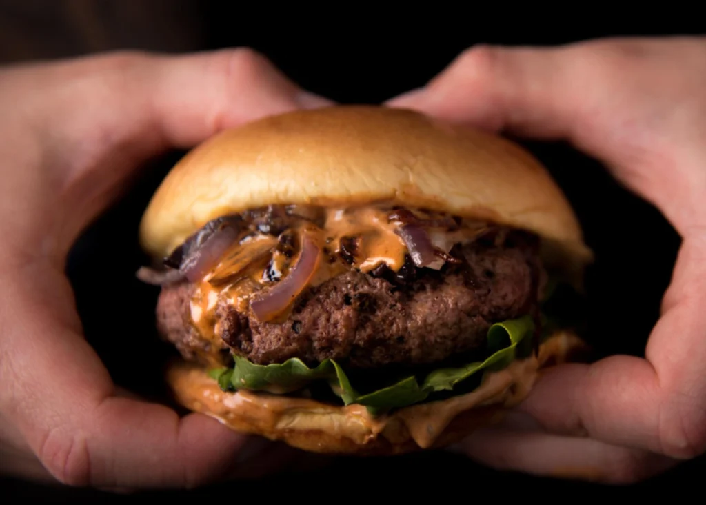 A sizzling cast-iron skillet on a rustic wooden surface, filled with juicy venison burger patties, their edges caramelized to a rich, golden-brown. Wisps of savory smoke curl upwards, mingling with the aroma of fresh herbs, garlic, and seared meat. In the background, a blend of natural light and warm, ambient lighting cast a cozy, inviting glow, highlighting the textural contrast between the rough-hewn wood and the smooth, glistening burgers. The composition is framed by the organic shapes of freshly chopped vegetables and a scattering of aromatic seasonings, creating a visually appetizing scene that captures the essence of "Cooking Methods for Venison Burgers". Venison minced meat