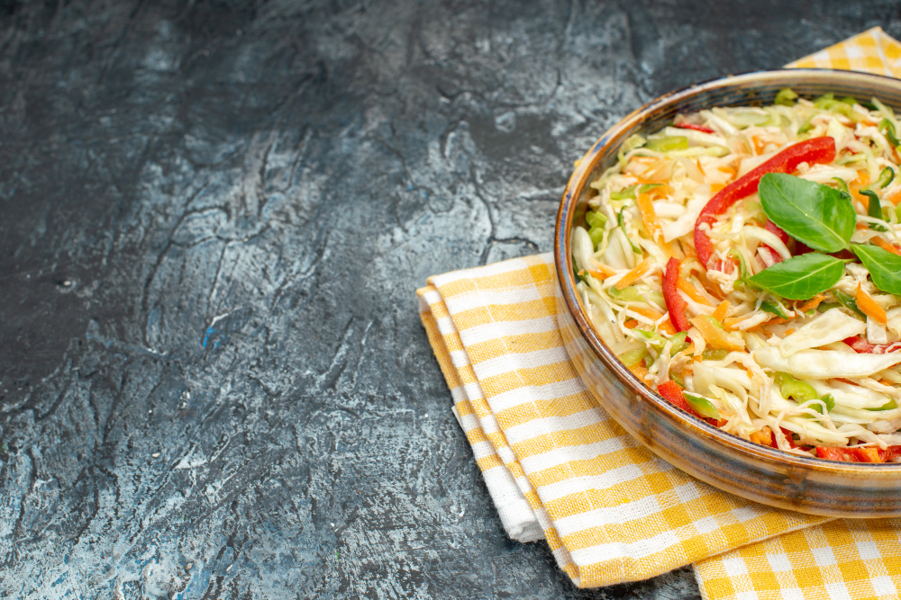 A wooden table with a bright white backdrop, the scene illuminated by natural, soft lighting. In the foreground, a large mixing bowl filled with crisp, shredded green and purple cabbage, sliced carrots, green onions, and crumbled ramen noodles. Beside the bowl, a chef's knife, a wooden cutting board, and a bottle of vinaigrette dressing. In the middle ground, a glass jar filled with sesame seeds and a small bowl of toasted slivered almonds. The background features a few simple kitchen utensils and a potted plant, creating a clean, minimalist composition that highlights the preparation of the ramen salad coleslaw.