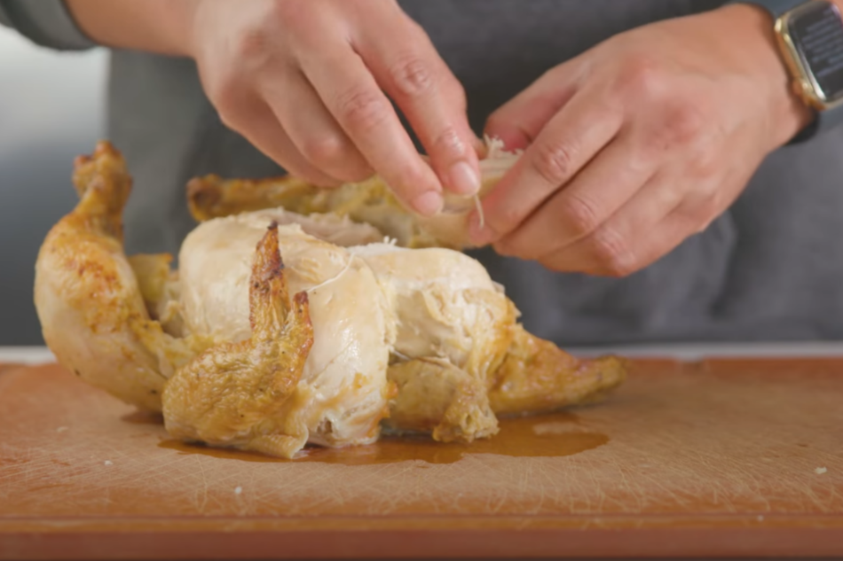 A close-up view of freshly cooked chicken breasts being shredded by hand on a wooden cutting board. The chicken is tender and juicy, with visible strands of meat separating easily. The lighting is warm and natural, casting gentle shadows that highlight the textural details. The background is blurred, putting the preparation process in sharp focus. The scene conveys a sense of culinary care and attention to detail, setting the stage for the creation of a delicious buffalo dip. Buffalo Chicken Dip