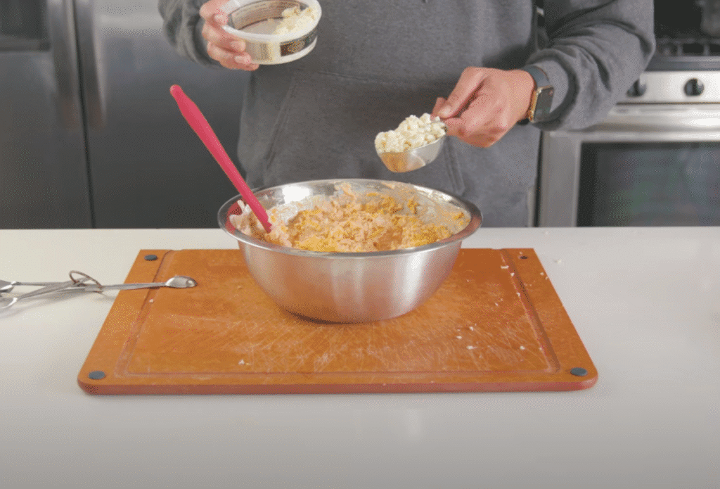 A pristine stainless steel mixing bowl sits on a clean, white countertop, filled with a creamy, golden-hued buffalo dip. The dip's consistency is smooth and pourable, with a glossy sheen. Nearby, a whisk and a spatula rest, suggesting the methodical blending of ingredients. The lighting is soft and diffused, accentuating the dip's inviting texture. The scene is captured from a slightly elevated angle, providing a clear view of the dip's perfect, velvety consistency, ready to be scooped and enjoyed.